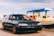 A black station wagon car is parked on a gravel surface near a container terminal. In the background, there are several colorful shipping containers stacked and a large blue and green gantry crane labeled 'INTERMOBIL'. The sky is partly cloudy.