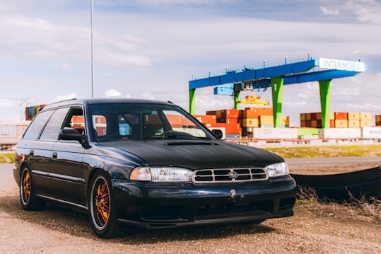 A black station wagon car is parked on a gravel surface near a container terminal. In the background, there are several colorful shipping containers stacked and a large blue and green gantry crane labeled 'INTERMOBIL'. The sky is partly cloudy.