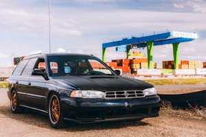 A black station wagon car is parked on a gravel surface near a container terminal. In the background, there are several colorful shipping containers stacked and a large blue and green gantry crane labeled 'INTERMOBIL'. The sky is partly cloudy.