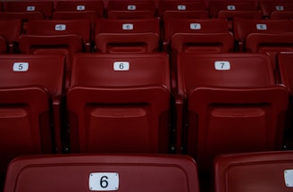 Red plastic seats arranged in rows, each marked with a number for identification. The seating appears uniform and is part of a larger auditorium or stadium setting.