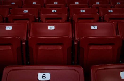Red plastic seats arranged in rows, each marked with a number for identification. The seating appears uniform and is part of a larger auditorium or stadium setting.
