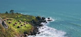 A scenic coastal view with a cluster of small, rustic cabins situated on a grassy cliff. The vibrant green grass contrasts with the rocky shoreline and the deep blue ocean waves crashing below. The ocean extends to the horizon, where it meets the sky, creating a sense of vastness.