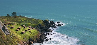 A scenic coastal view with a cluster of small, rustic cabins situated on a grassy cliff. The vibrant green grass contrasts with the rocky shoreline and the deep blue ocean waves crashing below. The ocean extends to the horizon, where it meets the sky, creating a sense of vastness.