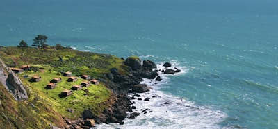 A scenic coastal view with a cluster of small, rustic cabins situated on a grassy cliff. The vibrant green grass contrasts with the rocky shoreline and the deep blue ocean waves crashing below. The ocean extends to the horizon, where it meets the sky, creating a sense of vastness.