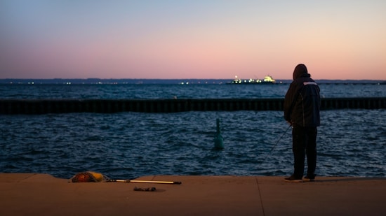 A person stands near the edge of a body of water, fishing quietly against the backdrop of a tranquil sunset. The distant horizon displays a faint glow from an offshore structure. Nearby, a fishing rod and bag lie on the ground.