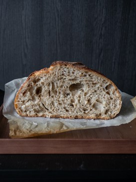 A rustic loaf of bread with a crunchy crust and a soft, airy crumb sits on parchment paper atop a wooden board. The background features a dark, textured surface that contrasts with the light color of the bread.