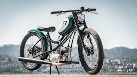 A vintage-style motorcycle is prominently displayed against an out-of-focus urban or mountainous landscape. The motorcycle has a sleek green and black design with number 77 painted on its fuel tank. It features a round headlight and spoked wheels, giving it a classic, retro aesthetic.