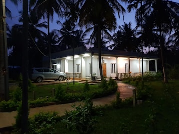 A modern single-story house surrounded by palm trees, situated in a lush garden setting. The house is illuminated from the inside, casting a warm glow that contrasts with the twilight sky. A car is parked adjacent to the house, and there is a pathway leading to the entrance, bordered by well-maintained greenery.