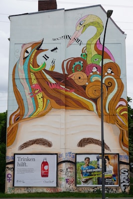 A large wall mural depicting a surreal scene with abstract animal forms, including a bird and a serpent-like creature. The artwork incorporates colorful, swirling patterns and elements such as eyes and text. Below the mural, there are two advertisements featuring text and images of a drink bottle and a smiling person. The wall itself is covered in graffiti, adding to the urban street art feel.