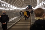 A tunnel with tiled walls is illuminated by a row of lights along the ceiling. Multiple people are walking and cycling through the tunnel, with a notable figure in the foreground wearing a mask. The tunnel extends into the distance, creating a sense of depth.