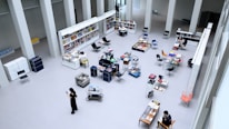 A spacious, open-plan library or bookstore with high ceilings and minimalist design. There are shelves filled with books, tables displaying a variety of items, and several chairs scattered throughout the space. People are seated or walking around, some engaged in reading or looking at their phones.