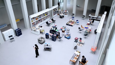 A spacious, open-plan library or bookstore with high ceilings and minimalist design. There are shelves filled with books, tables displaying a variety of items, and several chairs scattered throughout the space. People are seated or walking around, some engaged in reading or looking at their phones.