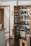 A well-organized kitchen pantry with wooden doors, featuring various items neatly arranged on shelves. There are jars of spices, baskets, bottles of oil, pasta, and canned goods. The door of the pantry has additional spice racks attached to it. The flooring is wooden, and there's a glimpse of stairs in the background.