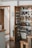 A well-organized kitchen pantry with wooden doors, featuring various items neatly arranged on shelves. There are jars of spices, baskets, bottles of oil, pasta, and canned goods. The door of the pantry has additional spice racks attached to it. The flooring is wooden, and there's a glimpse of stairs in the background.