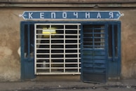 A small, rustic storefront with a blue framed window and door, featuring a sign above with Cyrillic letters. The window has bars and there's a partially open door. The building appears weathered with its faded beige facade.