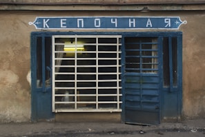 A small, rustic storefront with a blue framed window and door, featuring a sign above with Cyrillic letters. The window has bars and there's a partially open door. The building appears weathered with its faded beige facade.