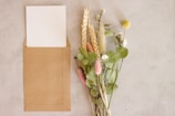 There is a brown envelope with a white card inside it placed on a light-colored surface next to a bouquet of various dried and fresh flowers.