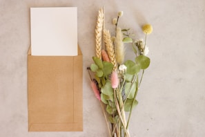 There is a brown envelope with a white card inside it placed on a light-colored surface next to a bouquet of various dried and fresh flowers.