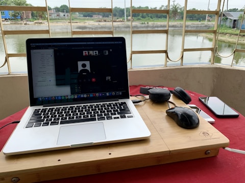 A laptop is on a wooden surface with a video conference on the screen. Beside the laptop, a pair of headphones, a mouse, a smartphone, and a charging cable are visible. The setup is placed near a large window with bars, overlooking a serene water body and some distant buildings and greenery.
