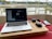 A laptop is on a wooden surface with a video conference on the screen. Beside the laptop, a pair of headphones, a mouse, a smartphone, and a charging cable are visible. The setup is placed near a large window with bars, overlooking a serene water body and some distant buildings and greenery.