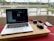 A laptop is on a wooden surface with a video conference on the screen. Beside the laptop, a pair of headphones, a mouse, a smartphone, and a charging cable are visible. The setup is placed near a large window with bars, overlooking a serene water body and some distant buildings and greenery.