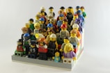 A collection of various LEGO minifigures arranged in rows, resembling an audience or group photo. The minifigures are diverse, depicting characters in different outfits and accessories, including helmets, glasses, and hats. They are set on a colorful LEGO base with blocks in shades of red, blue, green, and white.