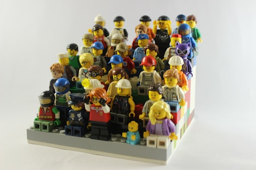 A collection of various LEGO minifigures arranged in rows, resembling an audience or group photo. The minifigures are diverse, depicting characters in different outfits and accessories, including helmets, glasses, and hats. They are set on a colorful LEGO base with blocks in shades of red, blue, green, and white.