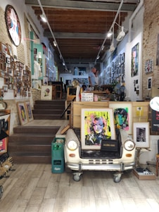 A cozy art shop adorned with numerous colorful paintings and photographs on the walls. The focal point is a vintage car front converted into a table displaying pop art-style portraits. The shop has a rustic feel with exposed brick walls and wooden beams on the ceiling. Two people are seen interacting in the background.