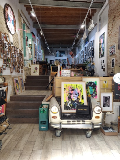 A cozy art shop adorned with numerous colorful paintings and photographs on the walls. The focal point is a vintage car front converted into a table displaying pop art-style portraits. The shop has a rustic feel with exposed brick walls and wooden beams on the ceiling. Two people are seen interacting in the background.