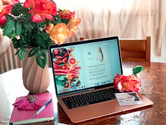 A MacBook sits open on a table displaying a document and an image with fresh fruits. Next to the laptop, there is an open notebook with a pen resting on it. A vase filled with vibrant pink and orange flowers decorates the table along with a single flower placed beside the laptop. An instant photo also lies nearby.