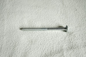 A metal screw lying horizontally against a textured, light-colored surface. The screw is silver in color and features a flat head and a threaded shaft.