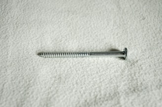A metal screw lying horizontally against a textured, light-colored surface. The screw is silver in color and features a flat head and a threaded shaft.