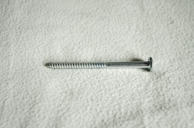 A metal screw lying horizontally against a textured, light-colored surface. The screw is silver in color and features a flat head and a threaded shaft.