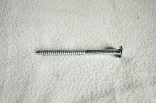 A metal screw lying horizontally against a textured, light-colored surface. The screw is silver in color and features a flat head and a threaded shaft.