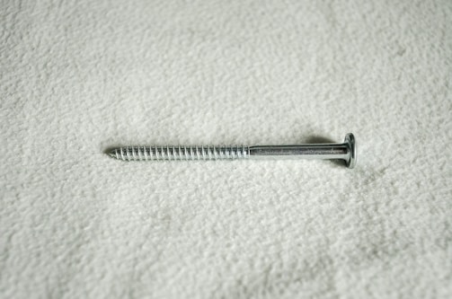 A metal screw lying horizontally against a textured, light-colored surface. The screw is silver in color and features a flat head and a threaded shaft.