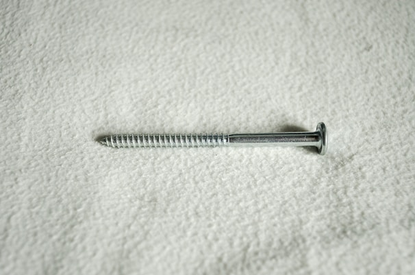 A metal screw lying horizontally against a textured, light-colored surface. The screw is silver in color and features a flat head and a threaded shaft.