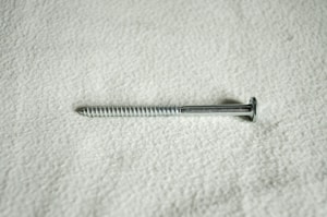 A metal screw lying horizontally against a textured, light-colored surface. The screw is silver in color and features a flat head and a threaded shaft.
