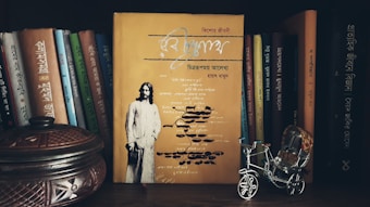 A collection of books with primarily Bengali titles is arranged on a bookshelf. In the foreground, there is a yellow book prominently displayed with an image of a man on the cover. To the side, a small, intricately designed metallic rickshaw and a decorative wooden container are visible.