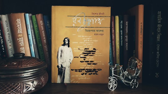 A collection of books with primarily Bengali titles is arranged on a bookshelf. In the foreground, there is a yellow book prominently displayed with an image of a man on the cover. To the side, a small, intricately designed metallic rickshaw and a decorative wooden container are visible.