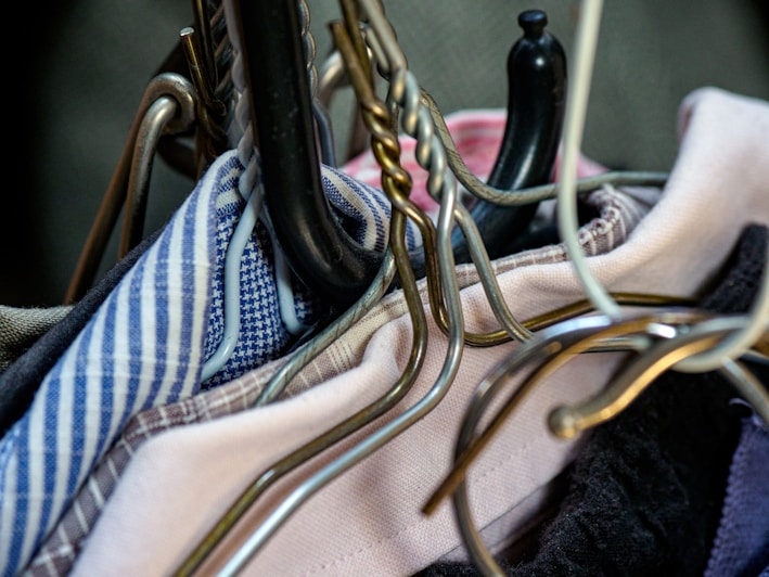 A close-up view of several clothes hangers entangled together, holding assorted garments. The hangers are made of various materials including metal and plastic, and the clothes display different patterns, such as stripes and checks. The fabric colors vary, adding a mix of textures and hues to the scene.