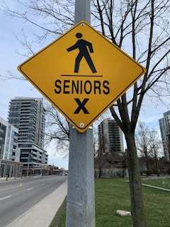 A yellow pedestrian warning sign indicating a seniors' crossing is mounted on a post along a city street. The area features high-rise buildings and leafless trees, suggesting an urban setting with a park or green space nearby.