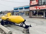 A bright yellow submarine is displayed on a trailer parked outside a brick building. The building has large signs advertising scuba services and lessons. Several adjacent shops are visible, including a pharmacy and a dry cleaner. The area appears to be a small shopping complex or strip mall.