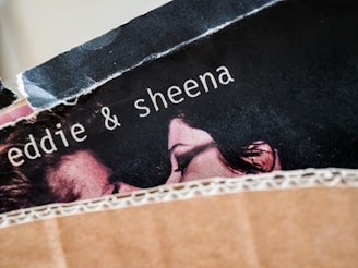 A close-up view of a torn poster featuring the names 'eddie & sheena' with part of a couple's faces visible. The image appears to be partially obscured by a piece of textured brown material, possibly cardboard.