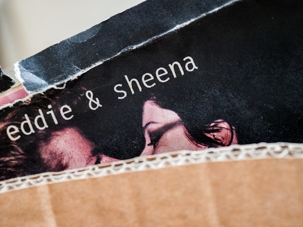 A close-up view of a torn poster featuring the names 'eddie & sheena' with part of a couple's faces visible. The image appears to be partially obscured by a piece of textured brown material, possibly cardboard.