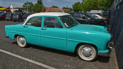 A vintage turquoise car with a white roof is parked in an outdoor parking lot. Surrounding the car are several modern vehicles, and the setting includes trees and a few commercial buildings in the background.