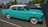 A vintage turquoise car with a white roof is parked in an outdoor parking lot. Surrounding the car are several modern vehicles, and the setting includes trees and a few commercial buildings in the background.