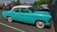 A vintage turquoise car with a white roof is parked in an outdoor parking lot. Surrounding the car are several modern vehicles, and the setting includes trees and a few commercial buildings in the background.