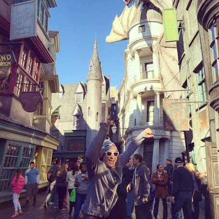 A lively street scene in a themed area resembling a magical or fantasy world, with people strolling and a prominent dragon sculpture displayed on a building. Multiple signs hang from the shops, and the architecture is whimsical, featuring turrets and Gothic elements.