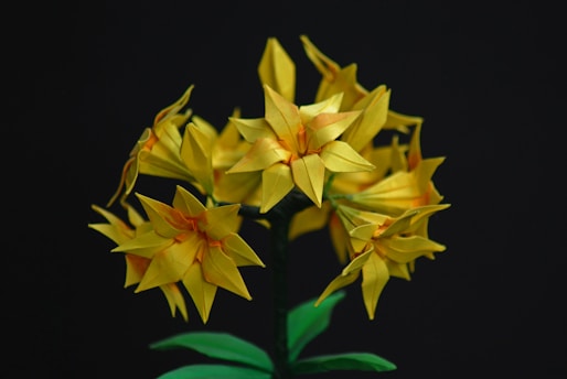 A cluster of origami flowers with intricate folds and vibrant yellow petals, supported by green paper stems and leaves, set against a dark background.