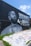 A large mural painted on a black wall features a monochrome portrait of a man with braided hair and a beard. The words 'Hustle & Motivate' appear in stylized lettering next to the portrait. The ground in front of the wall is covered with various colorful graffiti and signatures. Sparse patches of green grass can be seen along the edges.